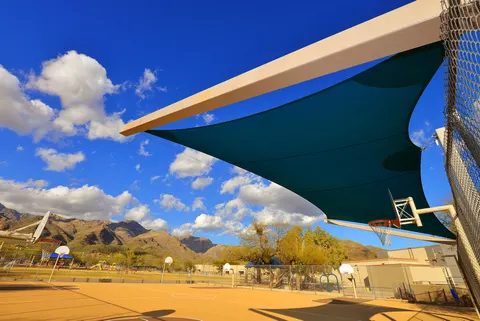 Upward view of a shade sail beside a basketball hoop.