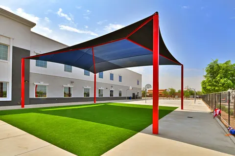 Dark shade canopy above a lawn beside a school building.
